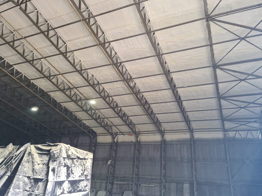 Spray foam applied to the underside of a large steel-framed hangar roof, covering sheets and purlins.