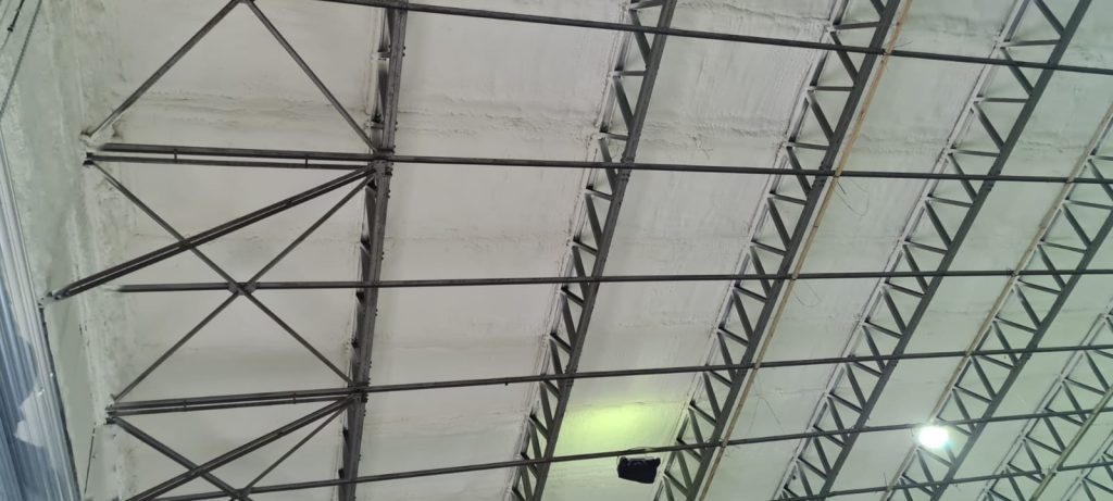 Spray foam insulation across the roof sheets between the trusses of a steel hangar.