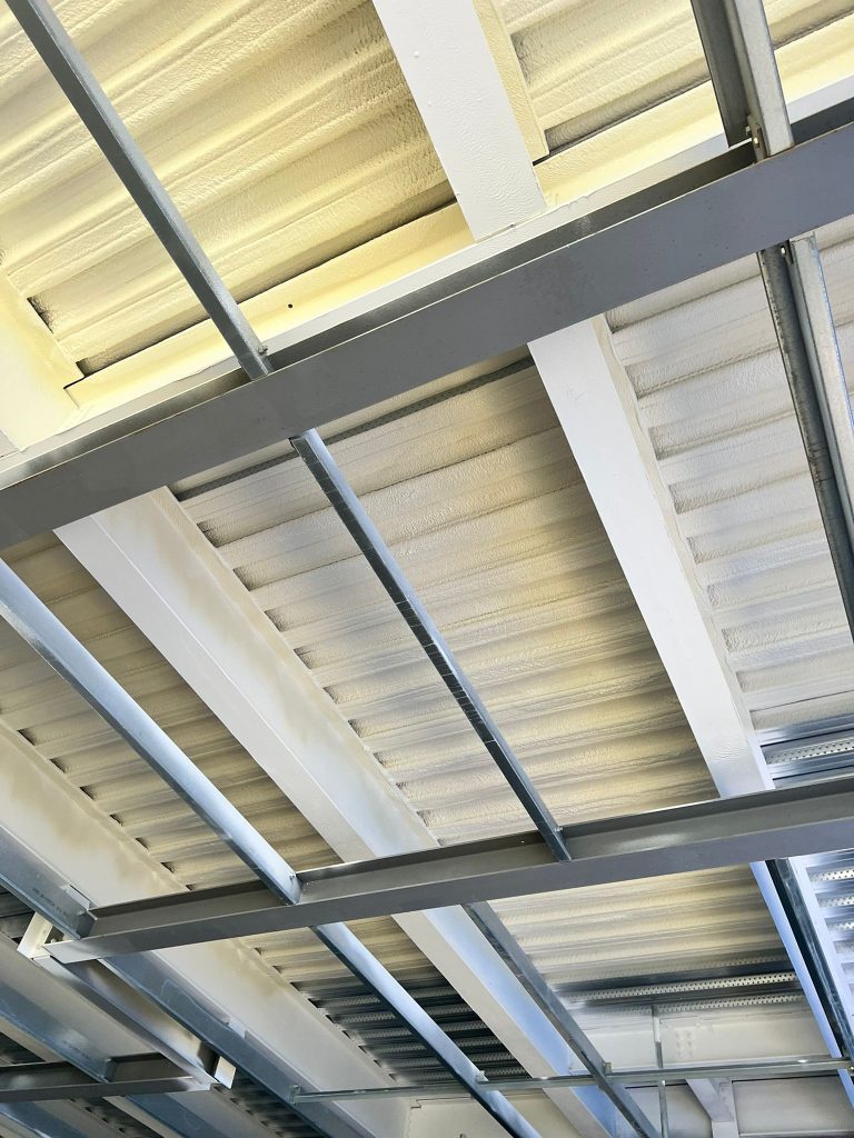 Nexseal LE closed-cell spray foam covering a profiled metal roof deck and the steel purlins below it.
